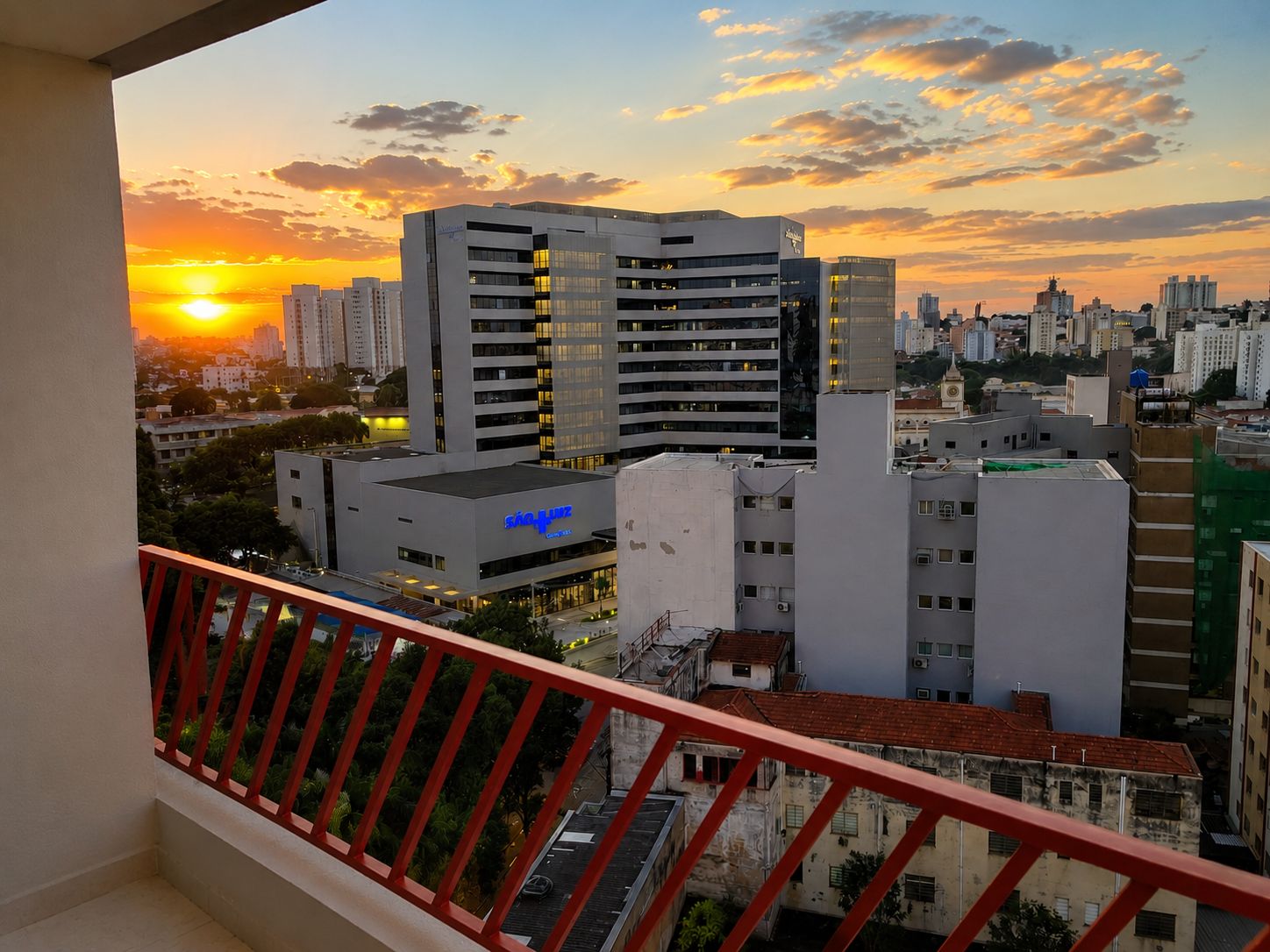 Un paisaje urbano con edificios modernos, un balcón con barandas rojas y un atardecer al fondo.
