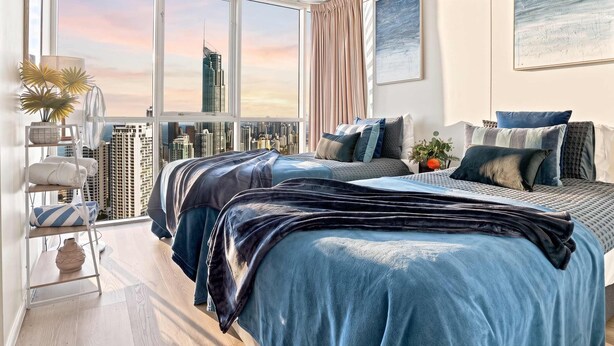 Un dormitorio con vistas a la ciudad, una cama con muchas almohadas, una mesita de noche y un ventanal grande.