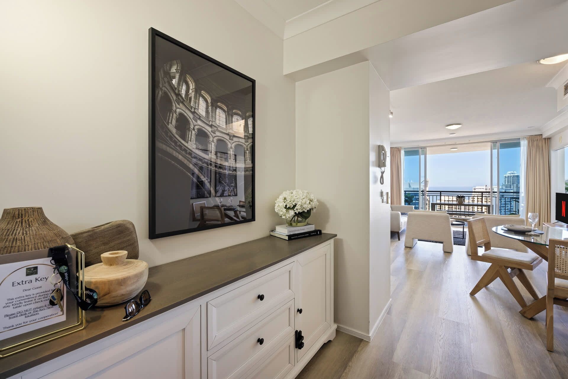 A hotel room with a large window, a dining area with a table and chairs, and a wall-mounted artwork.