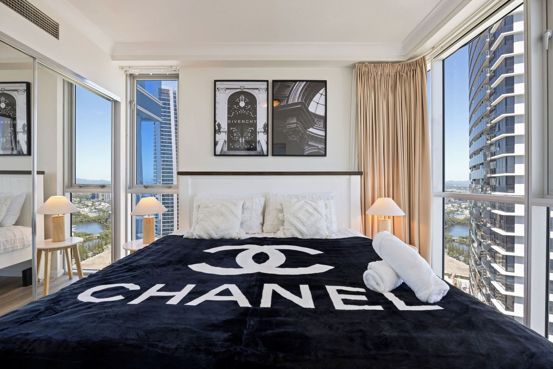 Een hotelkamer met een bed met een groot Chanel logo, twee ingelijste foto's aan de muur en een stadsgezicht door grote ramen.