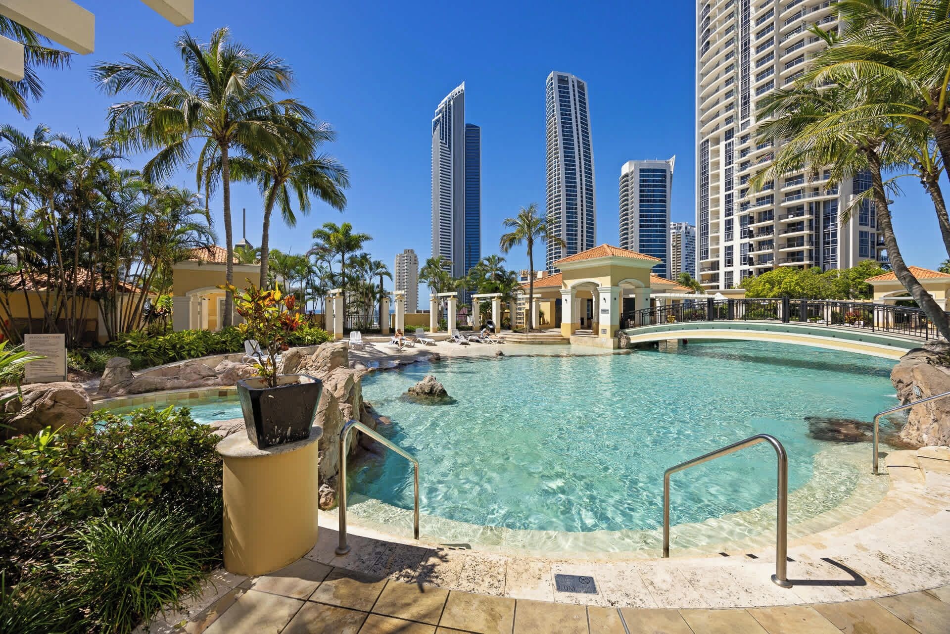 Ein Resort-Pool mit einer Brücke, Palmen und Hochhäusern im Hintergrund.