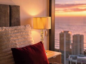 Una cama con una manta blanca con textura y una almohada roja, una lámpara de noche y una vista de la ciudad al atardecer a través de una ventana.