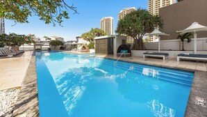 A swimming pool with lounge chairs and umbrellas, surrounded by tall buildings. - Hilton Surfers | Vogue on Orchid | 2BR Ocean View (Surfers Paradise)