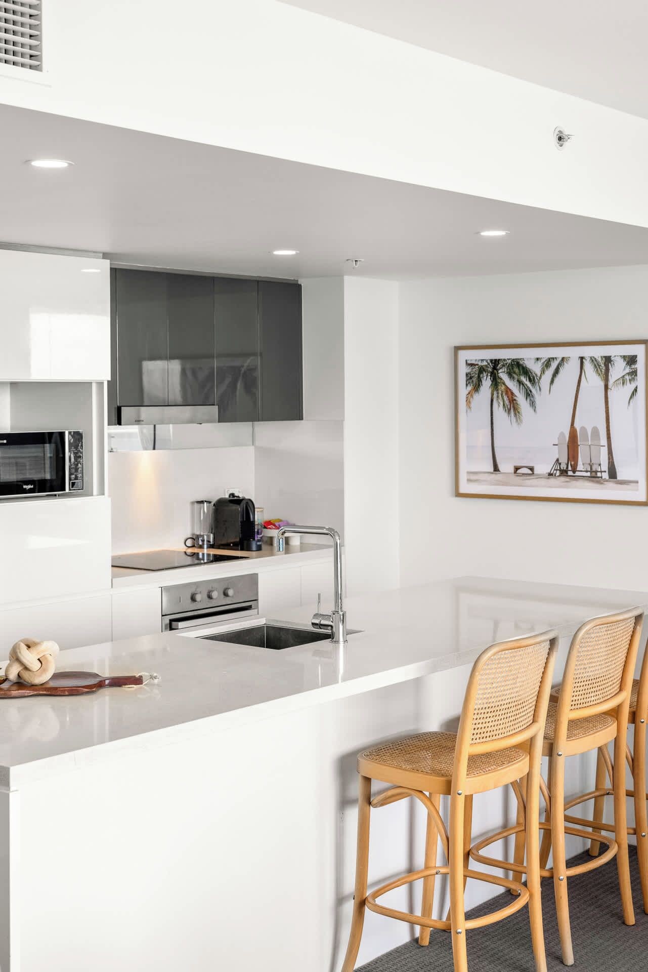 Una cucina moderna con mobili bianchi, un forno a microonde integrato e una grande isola con lavello. Ci sono due sedie in vimini all'isola.