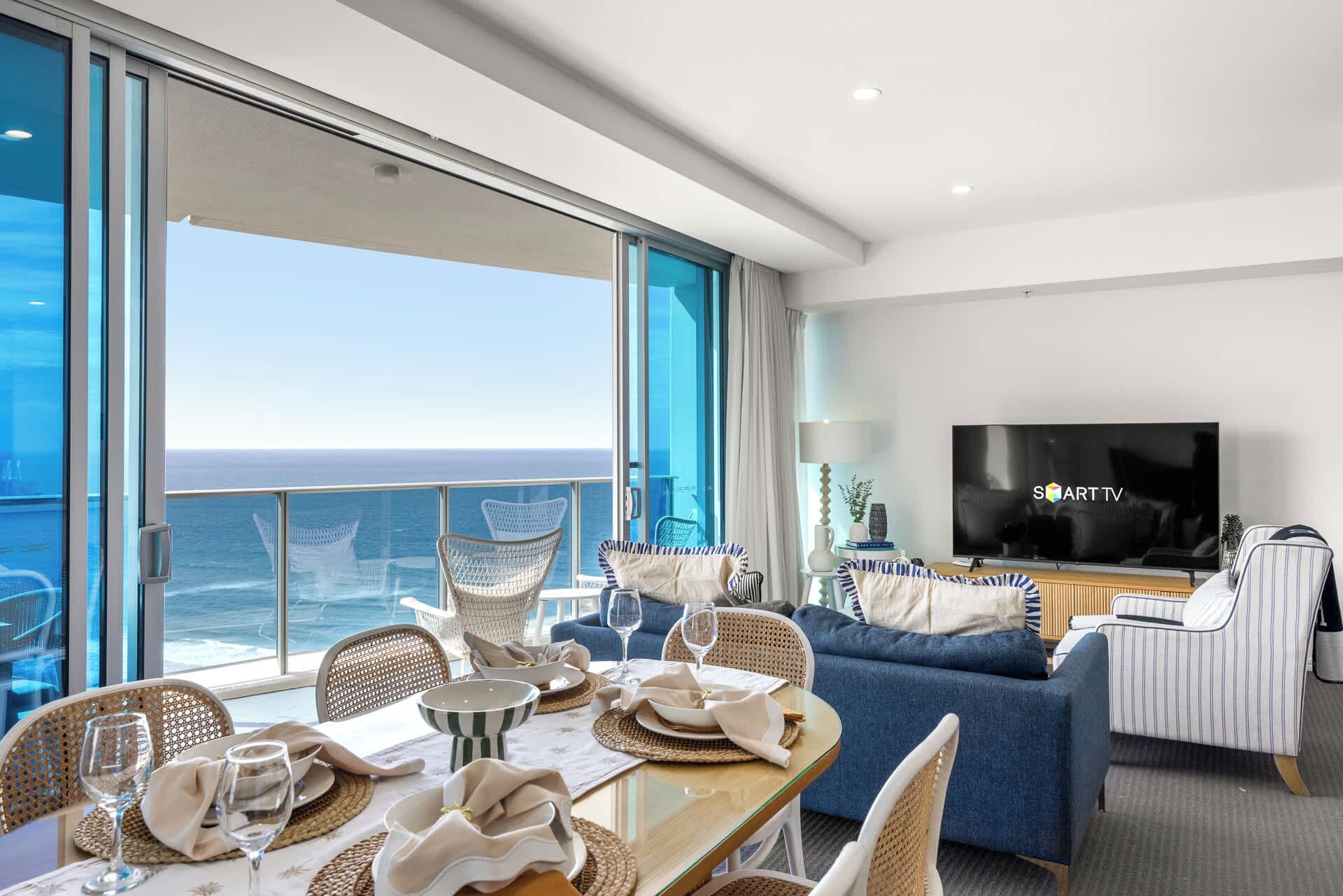 A modern living room with a large TV, comfortable seating, and a balcony with a view of the ocean.