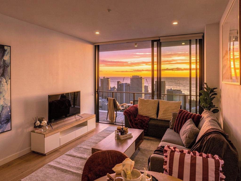 A modern living room with a large TV, comfortable seating, a coffee table, and a view of the city skyline at sunset.