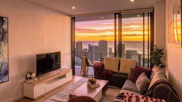 A modern living room with a large TV, comfortable seating, a coffee table, and a view of the city skyline at sunset.