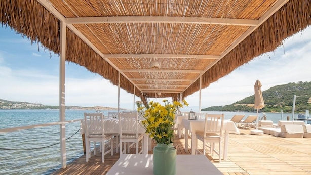 An outdoor dining area with a thatched roof, white chairs, and a table with a vase of yellow flowers. - Palmiye Beach & Rest (Bodrum)