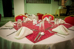 Una mesa de comedor dispuesta con servilletas blancas, cubiertos y copas de vidrio transparente, rodeada de sillas rojas con detalles amarillos.