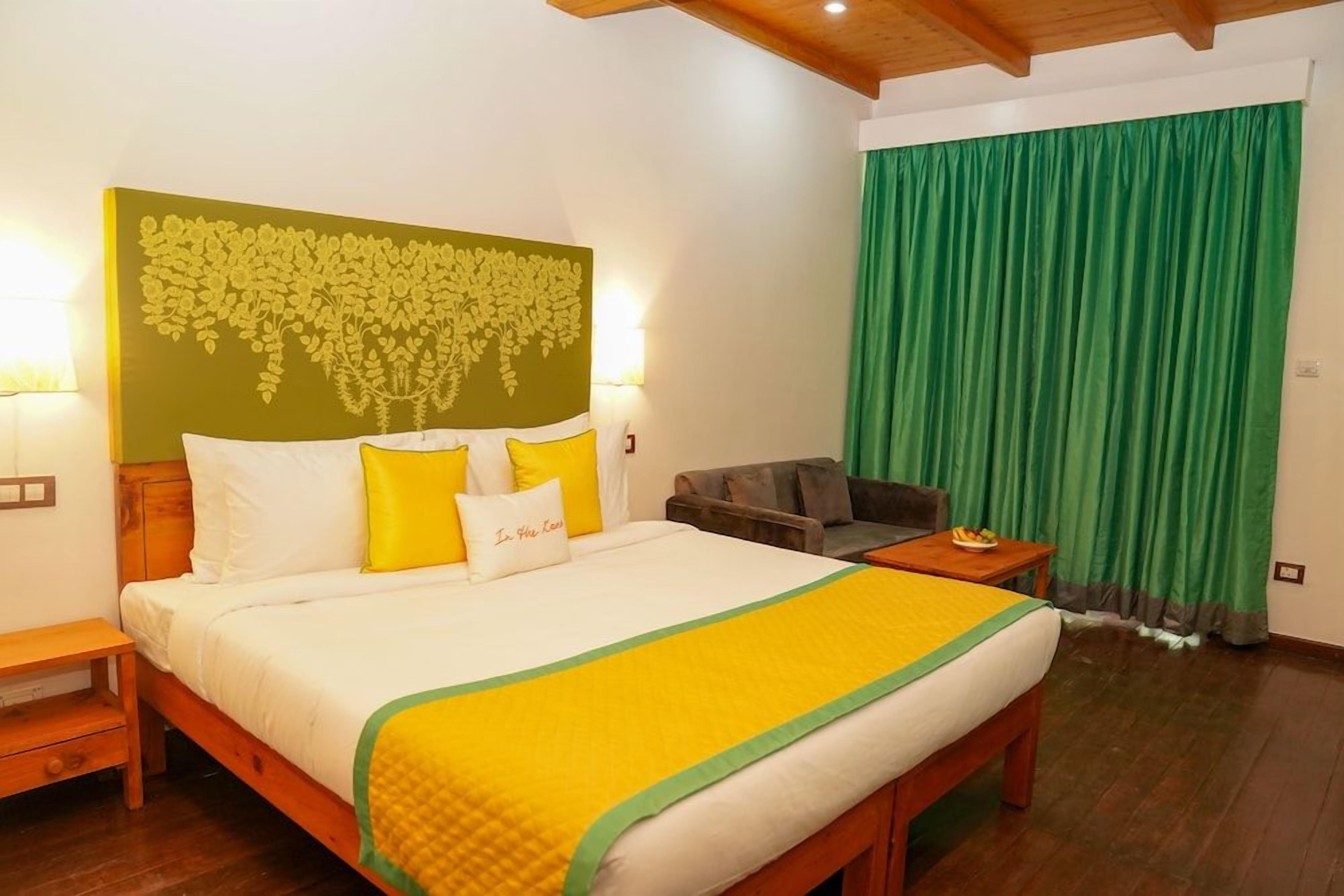 A hotel room with a large bed, a green curtain, a wooden headboard, and a small table with a fruit bowl.