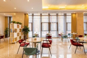 Un vestíbulo de hotel con mesas redondas, sillas rojas y verdes, una estantería con plantas y grandes ventanales con cortinas.