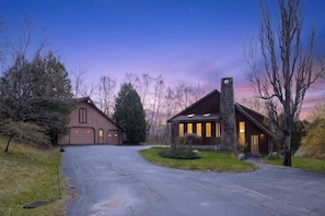 A two-story house with a driveway leading to a garage, surrounded by trees and a clear sky.