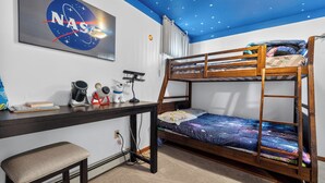 En våningssäng med ett sängöverkast med galaxmotiv, ett skrivbord med en dator, en stol, ett element och en NASA-affisch med ett rymduppdrag på väggen.