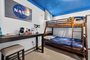 Una cama individual con edredón temático de galaxia, un escritorio con computadora, una silla, un radiador y un cartel de misión espacial de la NASA en la pared.