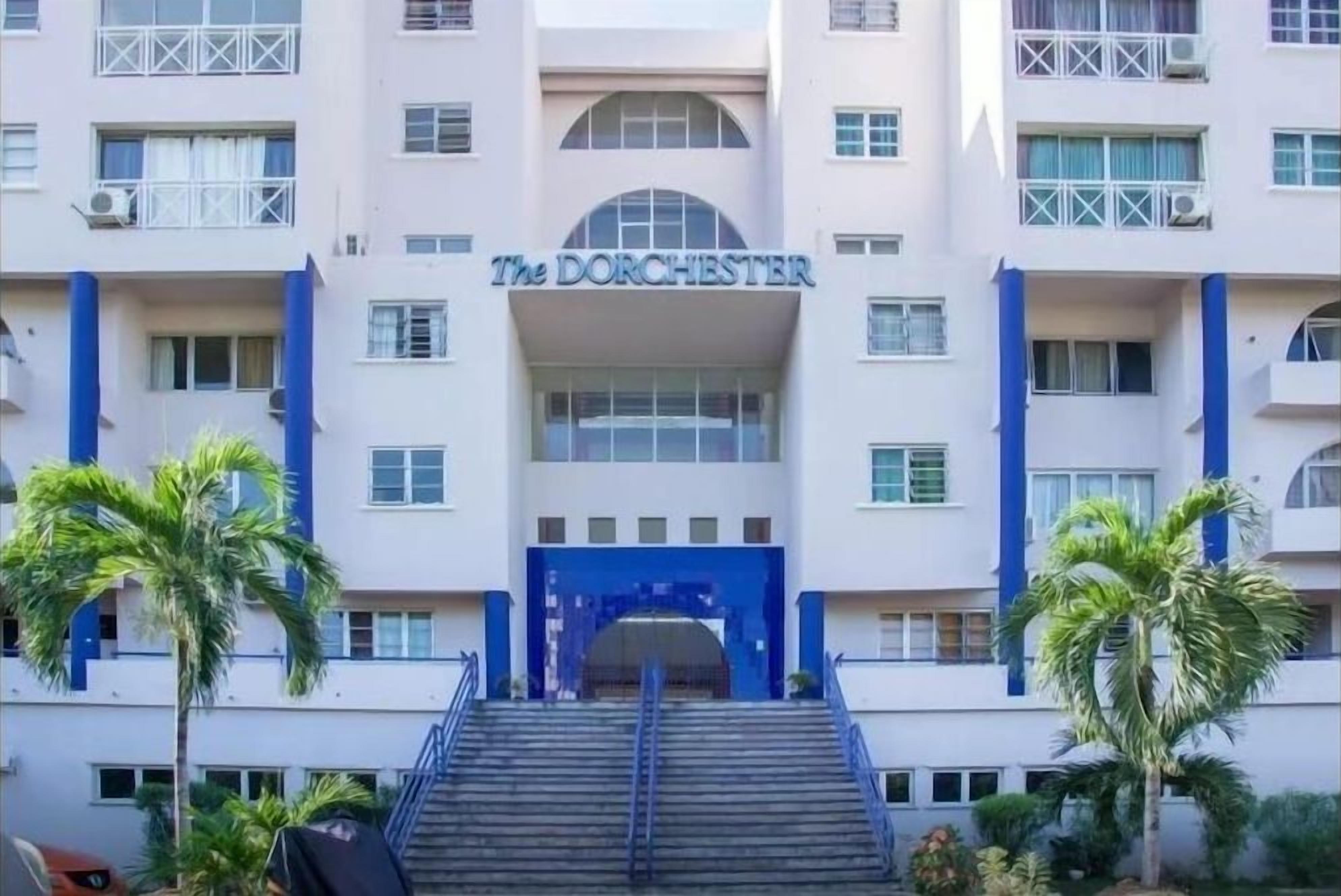 A hotel entrance with a prominent name sign, a staircase leading up to the entrance, and palm trees in the foreground.