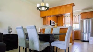 Uma sala de jantar com mesa preta e quatro cadeiras cinzentas, um candelabro, armários de madeira e uma geladeira de aço inoxidável.