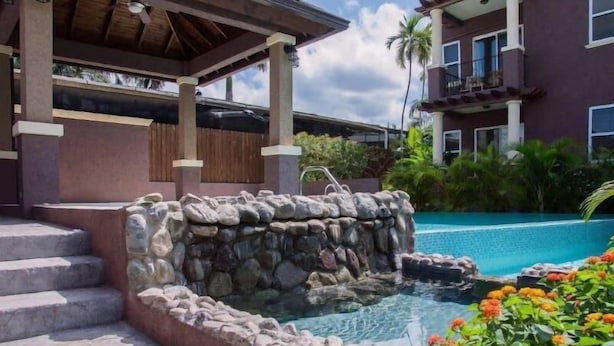 Banheira de pedra com degraus de acesso, rodeada por uma piscina e um edifício com varandas.