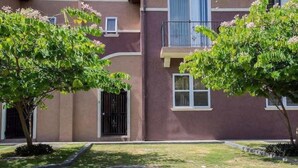 Um edifício residencial com estrutura de dois andares, varanda e pequena área de jardim com árvores e arbustos.