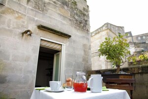 Un tavolo apparecchiato con una caraffa di bevanda rossa, una tazza, un piatto con pasticcini e una teiera, sistemato di fronte a un edificio con facciata in pietra.