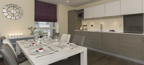 Una cocina moderna con microondas integrado, una mesa de comedor preparada para cuatro personas con platos de comida y vasos, y un reloj de pared.