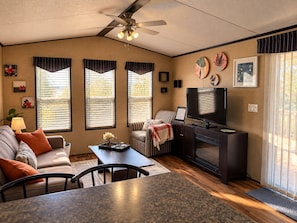 A well-lit living room with a ceiling fan, a television, and a fireplace. There are three windows with blinds, a sofa, and a dining table. - Peacedale | Luxury Cottage in Niagara-on-the-Lake • Pool • Pet-Friendly (Niagara-on-the-Lake)