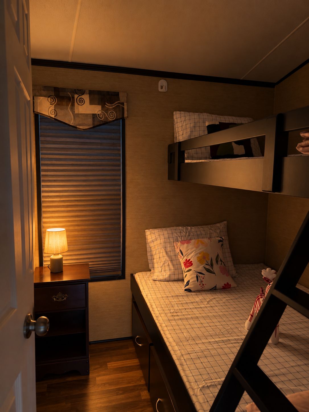 A compact bedroom with a bunk bed, a bedside table with a lamp, a window with blinds, and a decorative wall piece.