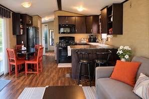 A compact kitchen with dark wood cabinets, a central island with stools, a dining area with red chairs, and a grey sofa with orange cushions. - Peacedale | Luxury Cottage in Niagara-on-the-Lake • Pool • Pet-Friendly (Niagara-on-the-Lake)