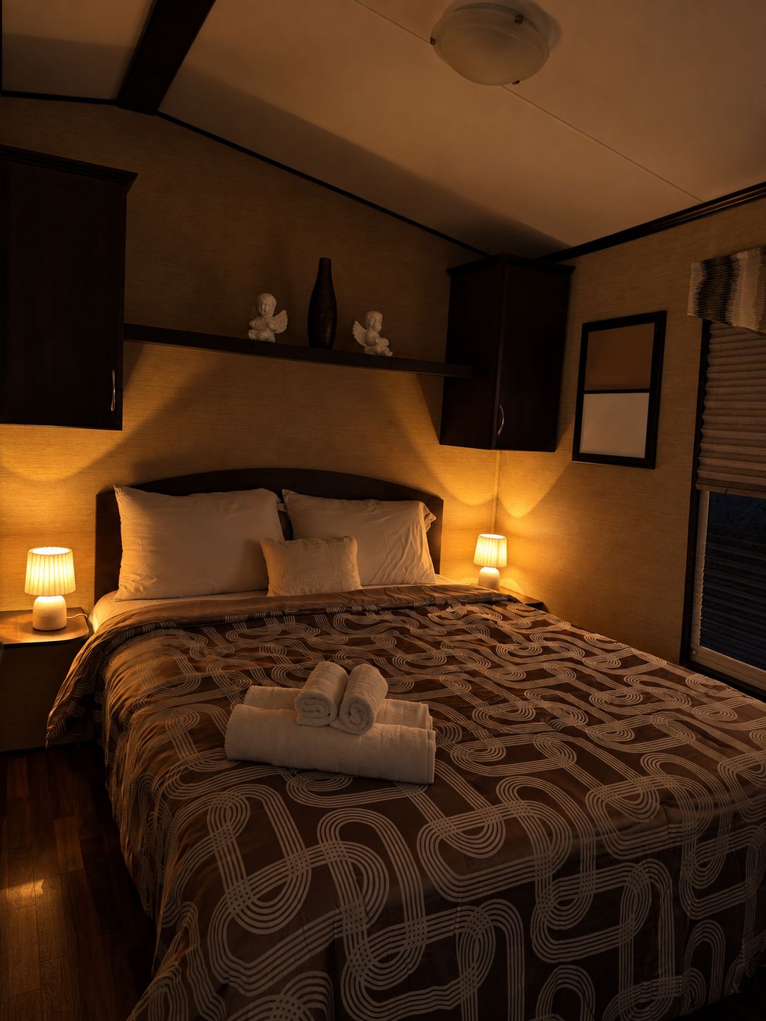 Habitación de hotel con una cama grande, dos lámparas de noche, una mesita, un cabecero, un estante en la pared con adornos y una ventana con persianas.