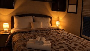Habitación de hotel con una cama grande, dos lámparas de noche, una mesita, un cabecero, un estante en la pared con adornos y una ventana con persianas.