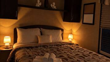 Habitación de hotel con una cama grande, dos lámparas de noche, una mesita, un cabecero, un estante en la pared con adornos y una ventana con persianas.