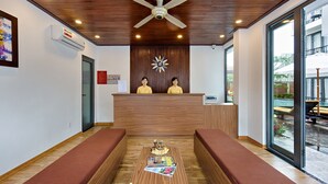 Zona de recepción de un hotel con techo de madera, mostrador de recepción, dos recepcionistas, un reloj y vistas a una piscina al aire libre.