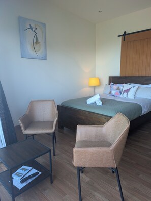 A hotel room with a bed, two armchairs, a table with a remote control, and a lamp. - The Cliffs Seaside Lodge (Papatotara)