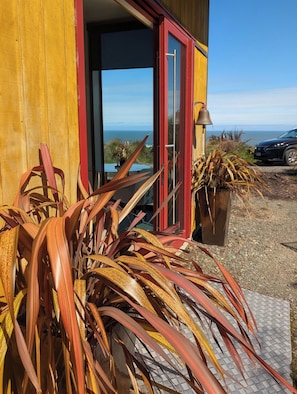Un edificio de madera con puerta roja, vista al océano y un coche estacionado afuera.