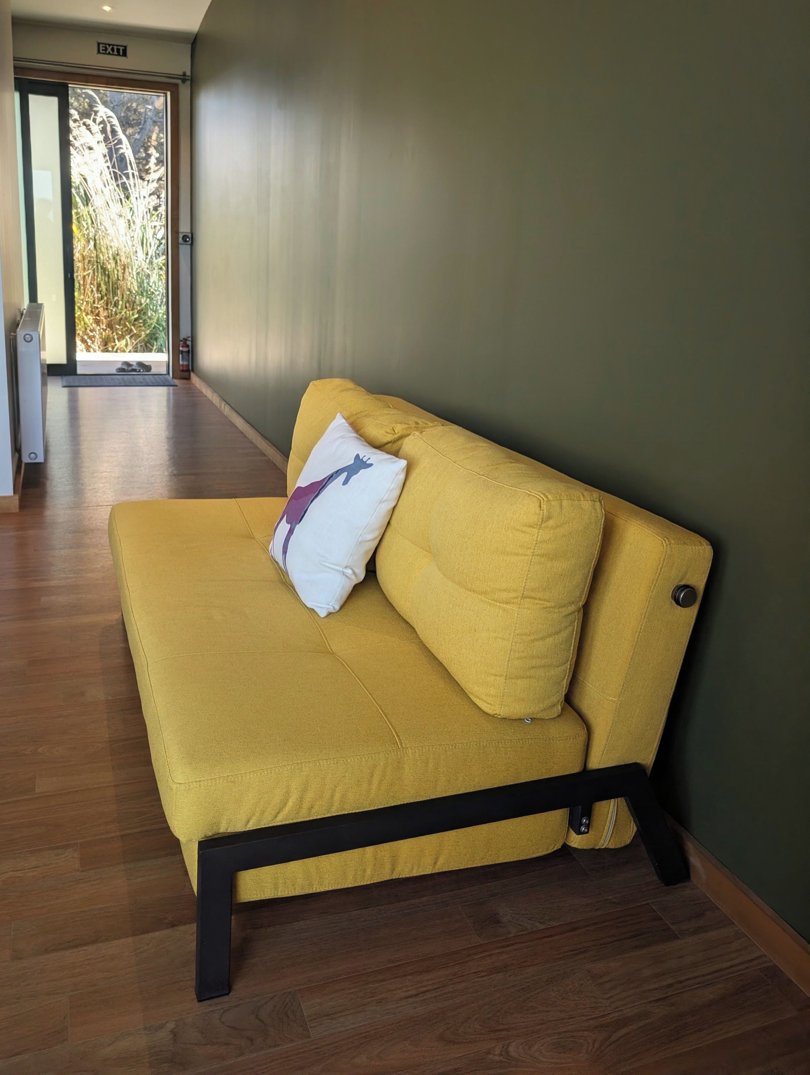 Un canapé jaune avec un seul coussin, adossé à un mur vert, sur un plancher en bois, et une porte de sortie visible au fond.