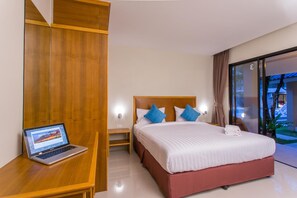 A modern hotel room with a wooden wardrobe, a bed with white bedding and blue pillows, a laptop on a wooden nightstand, and a view of the outdoors through a glass door. - Light Khao Lak Resort (Takua Pa)