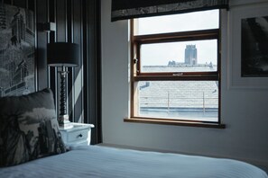 A bedroom with a bed, a nightstand, a lamp, and a window with a view of a building. - Posh Pads at The Casartelli (Liverpool)