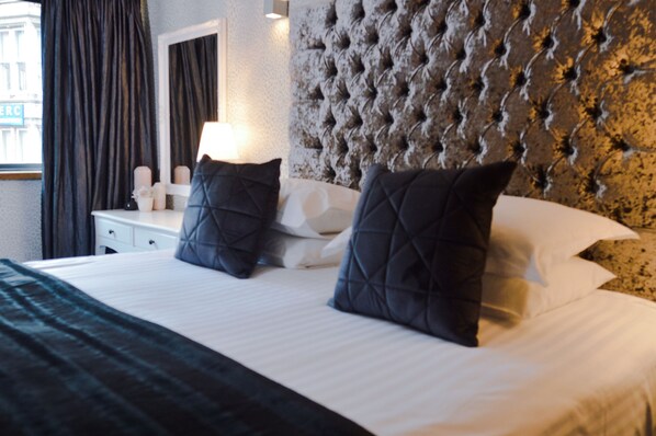A hotel room with a tufted headboard, a white bedside table with a lamp, and two dark pillows. - Posh Pads at The Casartelli (Liverpool)