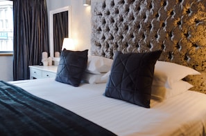 A hotel room with a tufted headboard, a white bedside table with a lamp, and two dark pillows. - Posh Pads at The Casartelli (Liverpool)