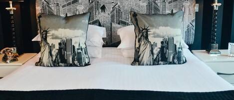 Habitación de hotel con una cama grande, dos almohadas con diseños de paisaje urbano, un papel tapiz con el horizonte de la ciudad y una mesita de noche con una lámpara.