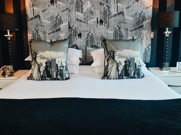 A hotel room with a large bed, two pillows with cityscape designs, a city skyline wallpaper, and a nightstand with a lamp.