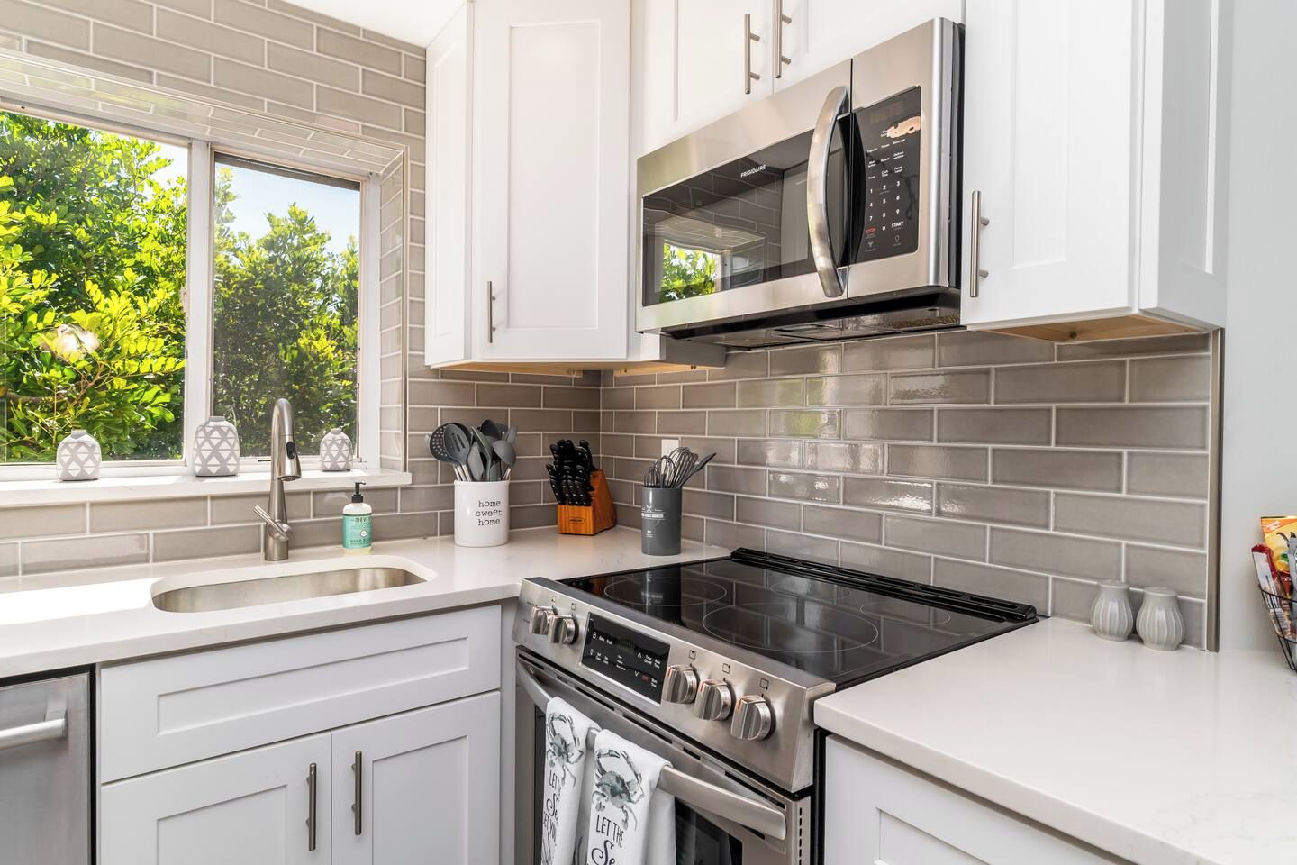 A modern kitchen with a stainless steel stove, microwave, and oven, white cabinetry, and a window with a view of greenery.