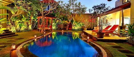 A swimming pool with a wooden deck, surrounded by plants and a gazebo.