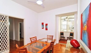 Un comedor con una mesa de vidrio y sillas de madera, un ventilador de techo y un cuadro abstracto rojo en la pared.