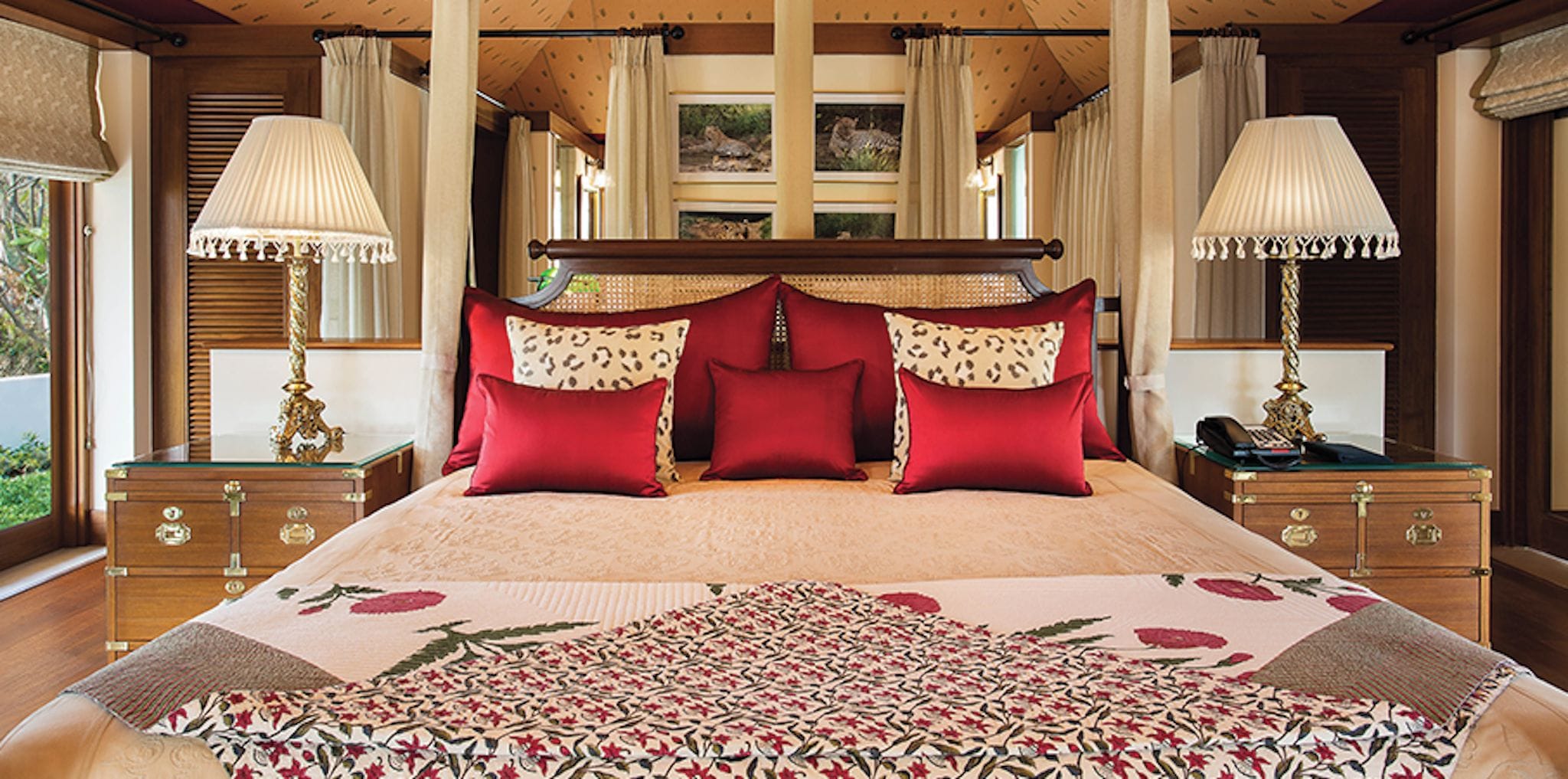 A large bed with a floral bedspread, multiple red and patterned pillows, and two bedside lamps.