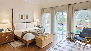 A hotel room with a four-poster bed, a bedside table with a lamp, a chair, a small table, and a view of the outdoors through large windows. - The Oberoi Sukhvilas Spa Resort, New Chandigarh (Kharar)
