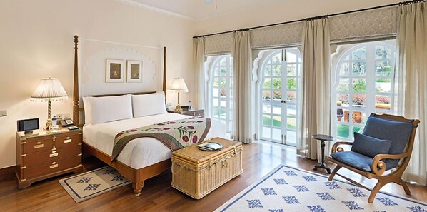 Habitación de hotel con cama con dosel, mesita de noche con lámpara, una silla, una pequeña mesa y vistas al exterior a través de grandes ventanales.