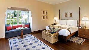 A bedroom with a bed, a sofa, a nightstand, a lamp, a dresser, and a window with a view. - The Oberoi Sukhvilas Spa Resort, New Chandigarh (Kharar)