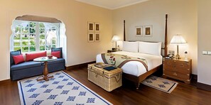 A bedroom with a bed, a sofa, a nightstand, a lamp, a dresser, and a window with a view. - The Oberoi Sukhvilas Spa Resort, New Chandigarh (Kharar)