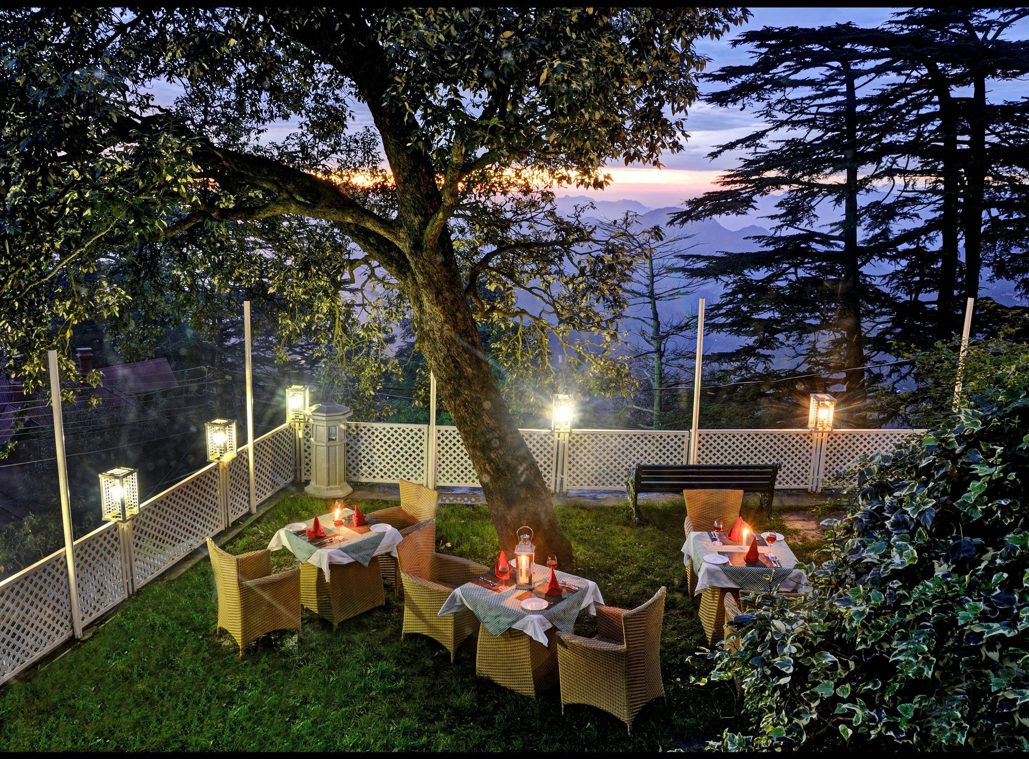 An outdoor dining area with wicker furniture and tables, surrounded by a white fence, with a view of mountains in the background.