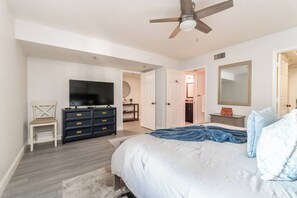 Una camera da letto con un letto, una cassettiera, una televisione, una ventola a soffitto e una porta che conduce al bagno.